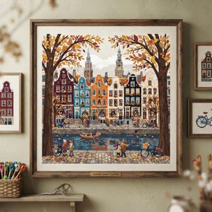 May include: A framed wall art print depicting an autumn scene in Amsterdam with colorful canal houses, a small wooden boat on the water, and people walking along the cobblestone path. Two large trees frame the view, and the text Amsterdam Autumn is visible on a small plaque at the bottom of the frame.