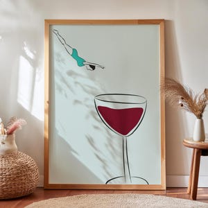 Può includere: Una stampa artistica incorniciata con un'illustrazione minimalista di un sub in costume da bagno turchese sopra un grande bicchiere di vino pieno di vino rosso. L'opera d'arte ha una cornice in legno chiaro ed è esposta su una parete bianca.