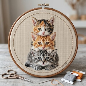 Pode incluir: Um bastidor de bordado circular de madeira contendo um design em ponto cruz de três gatinhos empilhados. O gatinho do topo é tricolor, o do meio é malhado laranja e o de baixo é malhado cinza. O texto M.S. 2024 está bordado no canto.