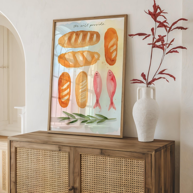 May include: A framed watercolour print depicting bread loaves and pink fish, with the text "He will provide" at the top. The artwork is displayed on a wooden cabinet, with a white vase and red plant to the right.