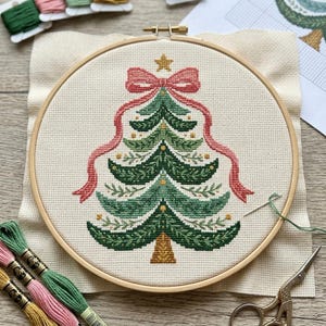May include: A cross-stitch project in a wooden embroidery hoop featuring a stylized Christmas tree design. The tree is stitched in shades of green with gold accents, topped with a gold star and a large pink bow with long flowing ribbons.