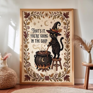 May include: A framed cross-stitch art piece featuring a black cat wearing a witch hat, standing on a stool and stirring a bubbling cauldron. The text reads, "THAT'S IT, YOU'RE GOING IN THE SOUP." The design is bordered by autumn leaves and flowers.
