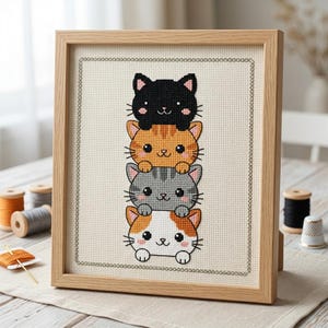 May include: A framed cross-stitch embroidery featuring a vertical stack of four cartoon cats. The cats are black, orange tabby, gray tabby, and calico, all with pink ears and whiskers. The design is stitched on cream fabric with a light wood frame.
