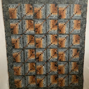 May include: A handmade quilt with a repeating pattern of square blocks. Each block features a log cabin design in shades of brown, gray, and orange. The quilt has a dark green border with a leaf pattern.