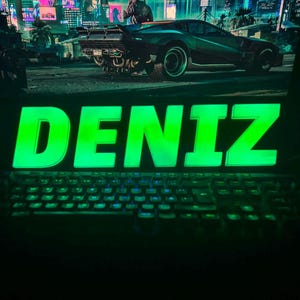 May include: A backlit green sign displaying the name "DENIZ" in bold, illuminated letters. A computer keyboard is in the foreground, with a monitor displaying a futuristic cityscape and a car in the background.