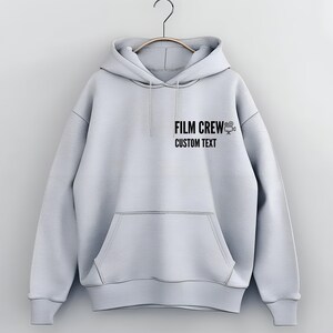 Pode incluir: Uma camisola de capuz cinzenta clara com um gráfico preto que diz "FILM CREW" e um ícone de câmara de filme. O texto abaixo do gráfico diz "CUSTOM TEXT".