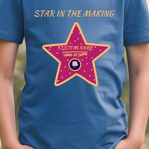 Könnte beinhalten: Ein blaues T-Shirt mit dem Text "STAR IN THE MAKING" und einem rosa Stern mit dem Text "CUSTOM NAME WALK OF FAME" und einem Filmkamera-Symbol.