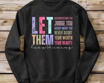 Op de afbeelding: Zwart sweatshirt met een kleurrijke afbeelding met de tekst "Laat ze je misverstaan, beoordelen, roddelen over je, twijfel nooit aan je waarde, je schoonheid. Blijf stralen."