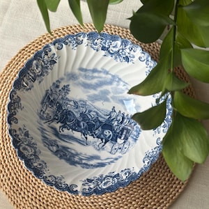 Puede incluir: Un cuenco decorativo blanco y azul con una ilustración detallada de un carruaje tirado por caballos. El cuenco tiene un borde festoneado y está colocado sobre un salvamanteles tejido. Hojas verdes en el fondo.