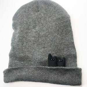 May include: A gray beanie hat with a folded brim. Two small black cat figures with button eyes are sewn onto the brim. The hat is made of a soft, knitted material.
