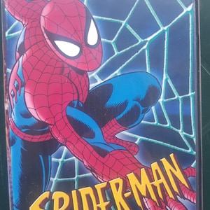Puede incluir: Una caja de DVD con Spider-Man en una pose dinámica. El superhéroe está representado con su clásico traje rojo y azul sobre un fondo azul con forma de telaraña. El título "SPIDER-MAN" está en letras amarillas. Texto: "L'intégrale 65 Episodes".