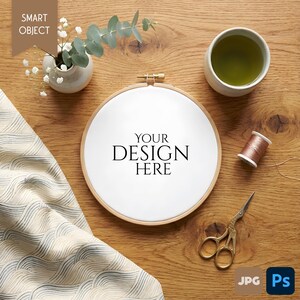 May include: A wooden embroidery hoop with the text "YOUR DESIGN HERE" is the focal point of this image. A small vase with white flowers and greenery, a cup of tea, and gold scissors are arranged on a wooden surface.