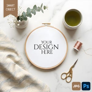 May include: A wooden embroidery hoop with the text "YOUR DESIGN HERE" is surrounded by craft supplies. A small vase with flowers, a cup of tea, a spool of thread, and gold scissors are also present.