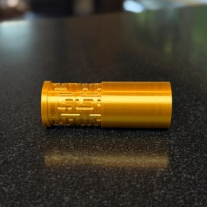 May include: A gold-colored metal cylinder with a patterned design. The cylinder is made of two pieces that fit together.