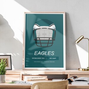 Może przedstawiać: Zielono-niebieski plakat z białym i czarnym kaskiem do futbolu amerykańskiego w środku. Tekst "EAGLES" znajduje się pod kaskiem w kolorze białym. Tekst "ESTABLISHED 1933" i "NFC EAST" znajduje się pod tekstem "EAGLES".