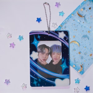 May include: A rectangular photo card featuring two people with dark and blue hair, framed by a blue and white starry design. The card has a silver chain attached at the top. Decorative stars and a blue fabric with gold moons are scattered around.