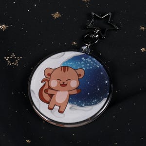 Könnte beinhalten: Ein runder Schlüsselanhänger mit einem Cartoon-Eichhörnchen auf einem Mond mit Sternenhimmel-Hintergrund. Der Schlüsselanhänger hat einen silbernen sternförmigen Verschluss und einen transparenten Kunststoffrand. Das Eichhörnchen ist braun mit rotem Schwanz und Wangen.