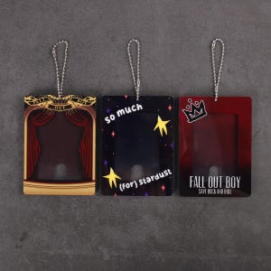 Könnte beinhalten: Drei rechteckige Schlüsselanhänger mit silbernen Ketten. Einer hat ein Bühnendesign mit dem Text "FALL OUT BOY". Ein anderer hat "so much (for) stardust" mit Stern-Grafiken. Der dritte hat eine Krone und den Text "FALL OUT BOY SAVE ROCK AND ROLL".