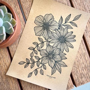 May include: A black ink botanical illustration of three flowers and leaves on tan paper. The artist's signature, "marygrace", is at the bottom. The artwork is displayed on a wooden surface. The drawing is detailed and intricate.