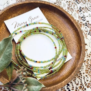 May include: A collection of beaded waist beads in a wooden bowl. The beads are mainly light green with red, blue, and gold accents. A white card with the text "Mary Grace Jewellery" is also in the bowl.