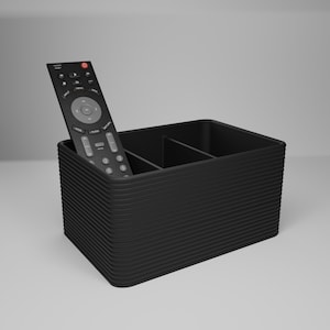 May include: A black, rectangular remote control organizer with three compartments. A black remote control with gray buttons is placed inside the organizer. The organizer has a ridged, curved design.