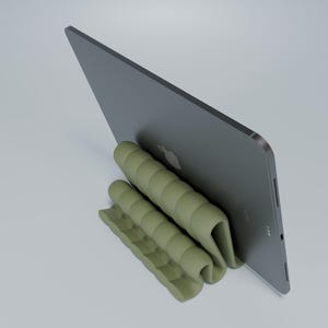 May include: A green, flexible, and foldable stand for a tablet or laptop. The stand is made of a soft, rubber-like material and has a series of ridges that create a stable base for the device. The stand is designed to prop up the device at an angle, making it easier to view and use.