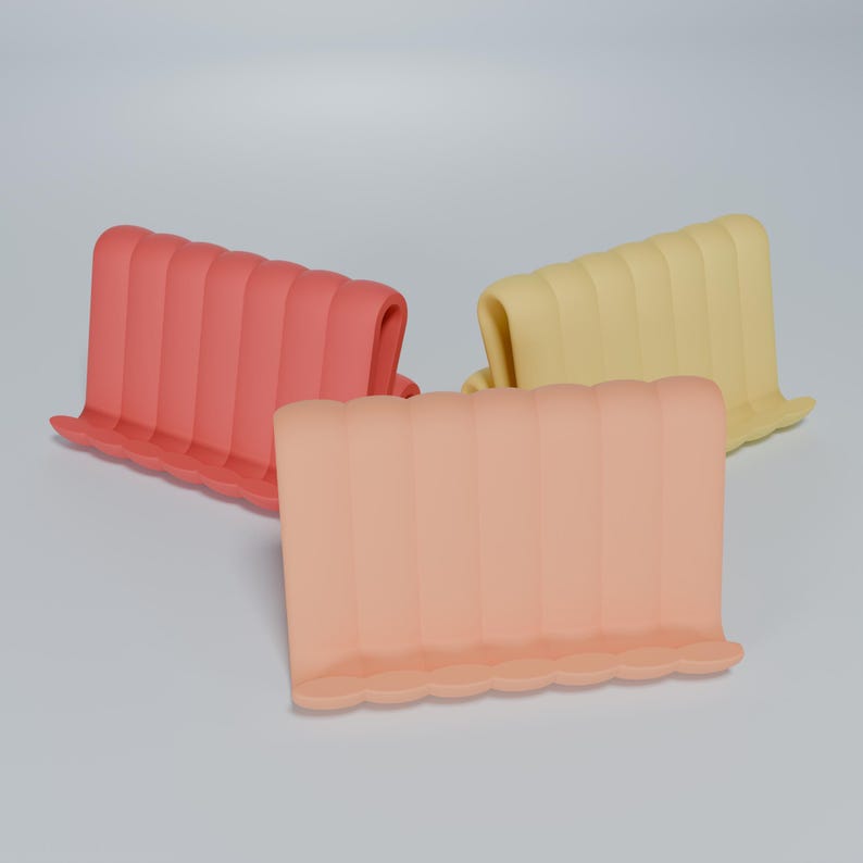 May include: Three silicone phone stands in different colors: pink, peach, and yellow. Each stand has a wavy design and is shaped like a rectangle.