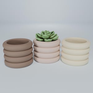 May include: Three ceramic planters with a ribbed design. The planters are in brown, pink, and cream. A small green succulent plant is in the pink planter.