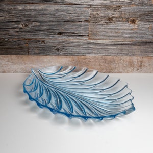 May include: A light blue, leaf-shaped glass serving dish. The dish has a detailed, textured design with curved lines and a scalloped edge. The dish is approximately 38 cm long.