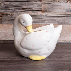 May include: A white ceramic goose-shaped planter with a yellow beak and base, and small red dots. The goose is in a curved pose, with its head near its body. The planter is on a dark wooden surface, with a wooden plank background.