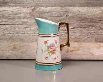 Może przedstawiać: Wazon ceramiczny w stylu vintage z turkusową krawędzią i uchwytem oraz złotymi akcentami. Korpus wazonu jest biały z kwiatowym wzorem w kolorze różowym, żółtym i niebieskim. Uchwyt jest złoty.