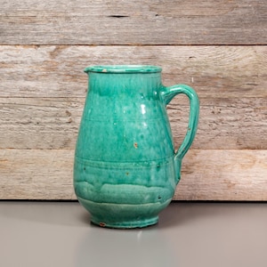 May include: A green ceramic jug with a rounded body and a handle. The jug has a rustic, vintage look.