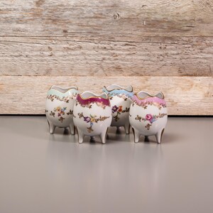 Peut inclure: Quatre petits pots à œufs en porcelaine avec des motifs floraux dans des tons de rose, bleu et vert. Chaque pot a trois petits pieds.