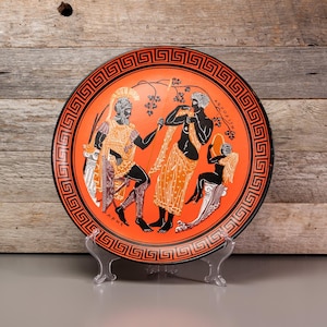 May include: A red ceramic plate with a black and orange design depicting a scene from Greek mythology. The plate features a Greek key border and the words "APHRODITE" and "ARES" in black.