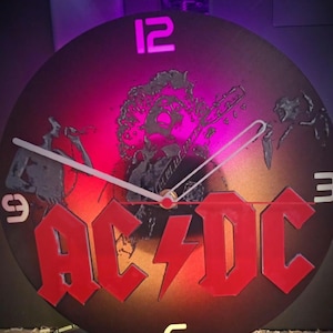 Könnte beinhalten: Eine schwarze Schallplattenuhr mit einem roten und orangefarbenen Verlaufshintergrund. Das Zifferblatt der Uhr zeigt das AC/DC-Logo und eine Silhouette einer Person, die Gitarre spielt. Die Zahlen auf dem Zifferblatt sind weiß.