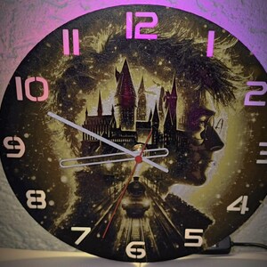 Puede incluir: Reloj de pared redondo con la silueta de una persona con gafas y un diseño de castillo. La esfera del reloj tiene un fondo oscuro con números y manecillas rosas iluminadas. Un tren es visible en las vías. Es un artículo decorativo.