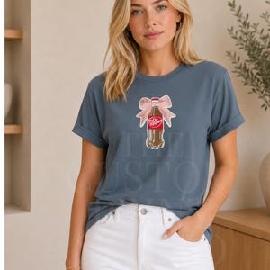 Peut inclure: T-shirt bleu ardoise avec un motif de bouteille de Dr. Pepper orné d'un nœud rose. Le t-shirt a des manches retroussées et est porté avec un jean blanc. Le texte "THE CUSTOM" est partiellement visible sur le t-shirt.