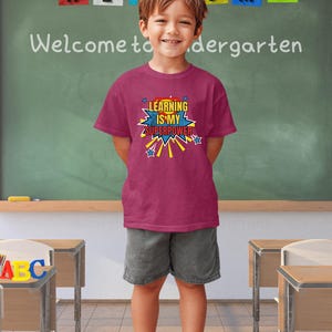 Puede incluir: Un niño lleva una camiseta de color bayas con el texto "LEARNING IS MY SUPERPOWER" en un estilo de cómic. El niño está de pie en un aula, con una pizarra y escritorios en el fondo.