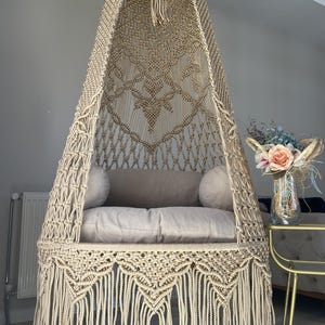 Puede incluir: Sillón colgante de macramé beige con la parte superior redondeada y flecos. La silla presenta un diseño tejido con un asiento acolchado y dos cojines. Un arreglo floral decorativo está cerca.