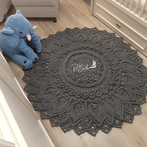 May include: A gray crocheted round rug with a mandala design. The rug is on a wooden floor in a room with white walls.