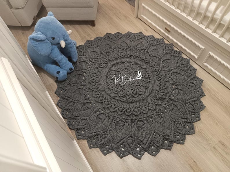 May include: A gray crocheted round rug with a mandala design. The rug is on a wooden floor in a room with white walls.