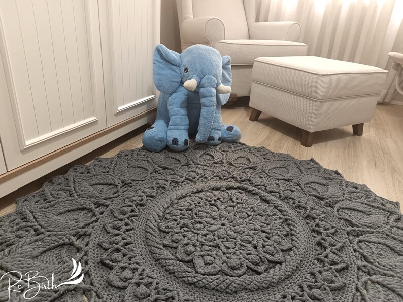 May include: A gray crocheted rug with a large circular pattern in the center. A blue plush elephant toy is sitting on the rug.
