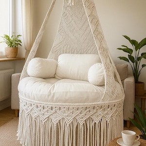 May include: A cream macrame hanging chair with a round seat and fringe. The chair has a canopy and is suspended from the ceiling. Two cylindrical pillows and a large cushion are on the seat. The chair is in a living room setting, with a neutral colour scheme.