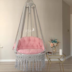Peut inclure: Un fauteuil suspendu en macramé gris avec un coussin rose et des franges. Le fauteuil est suspendu au plafond dans une pièce aux murs blancs et un sol en bois.