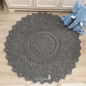May include: A gray crocheted round rug with a mandala design. The rug has a delicate, intricate pattern with a central flower motif.
