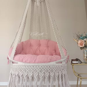 Pode incluir: Uma cadeira suspensa de macramé branca com um almofada rosa e acabamento em franjas. A cadeira está pendurada em um teto branco.