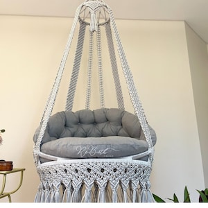 May include: A white macrame hanging chair with a gray cushion. The chair has a fringe border and is suspended from the ceiling.