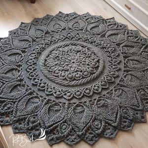 May include: A gray crocheted rug with a large circular mandala design. The rug has a detailed pattern with intricate details and a textured surface.