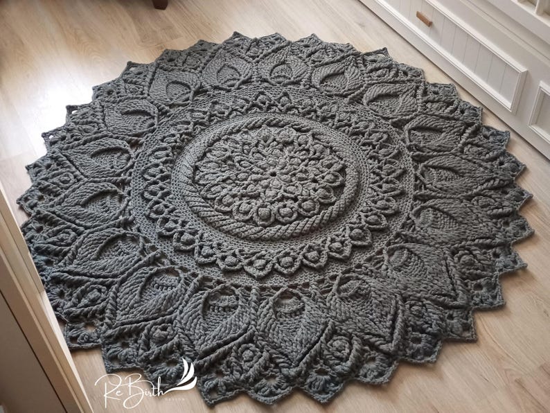 May include: A gray crocheted rug with a large circular mandala design. The rug has a detailed pattern with intricate details and a textured surface.