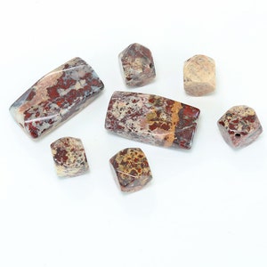 May include: A collection of seven jasper beads in red, brown, and white. The beads are in various shapes, including rectangular and cube shapes. The beads are polished and have a smooth surface.